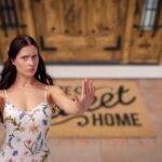 in primo piano una donna che dice no e come sfondo una porta d'ingresso con uno zerbino